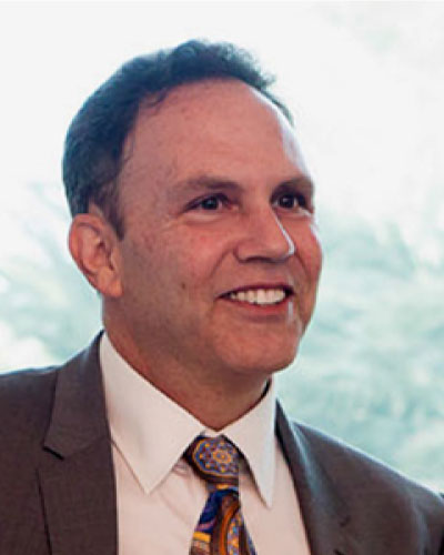 Smiling man in a brown suit and white shirt with a patterned tie, looking slightly to the right.