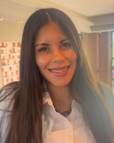 Portrait of a smiling woman with long dark hair, wearing a white blouse and beaded necklace, indoors with warm lighting.