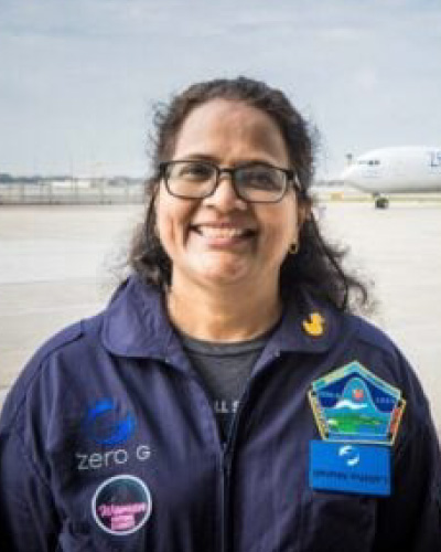 Smiling woman in a blue flight jacket with mission patches, standing on an airport tarmac with a plane in the background.