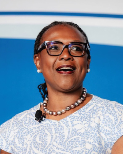 Woman wearing glasses and a pearl necklace speaks on stage, with a lapel microphone attached to her dress poster.