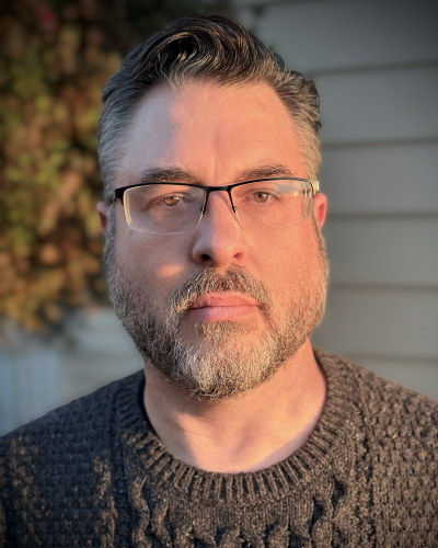 Michael Laine | ISDC 2026 Man with graying hair and beard wearing glasses, looking at the camera in a dark knit sweater outdoors, with a house and foliage in the background.