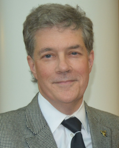Smiling middle-aged man with gray hair in a checkered blazer, white shirt, and black tie, facing the camera for a professional headshot.