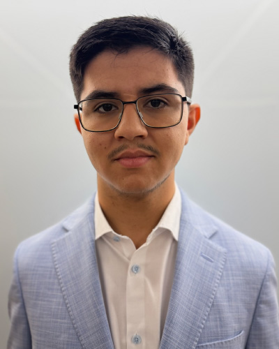 Young man wearing a light blue blazer and white shirt with glasses, looking at the camera in a neutral pose.
