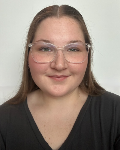 Young woman with clear-framed glasses smiling at the camera, wearing a black top against a light neutral background.
