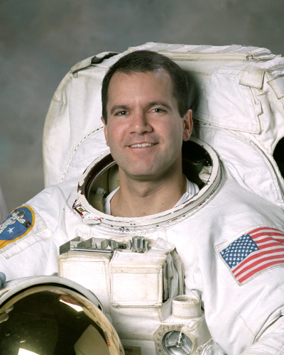 Astronaut in a white spacesuit smiling, with a U.S. flag patch on the arm and a mission patch on the other shoulder.
