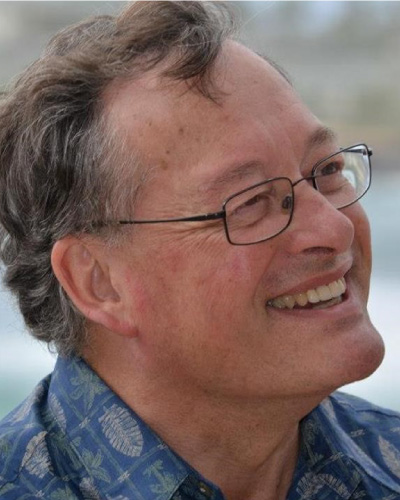 Profile of a smiling man with glasses and gray hair, wearing a blue patterned shirt outdoors.