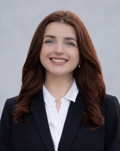Portrait of a smiling woman with shoulder-length brown hair, wearing a black blazer and white shirt, against a neutral gray backdrop.