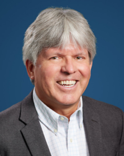 Mark-A-Skinner | ISDC Smiling man in a dark blazer and light shirt posing against a solid blue background.