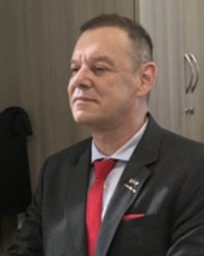 Michael-Dean-Puckett | ISDC Middle-aged man in a dark suit and red tie sits in an office, looking to the left, with a lapel pin visible on his jacket.