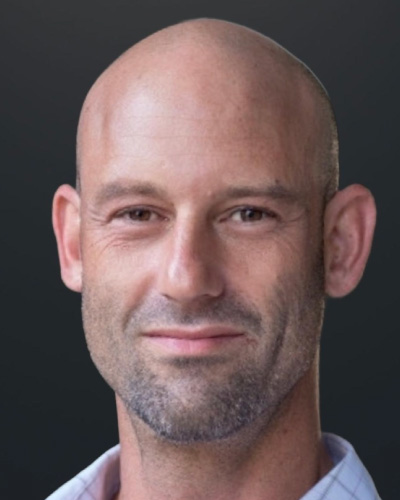 Close-up portrait of a bald man with light stubble, smiling slightly, against a dark gray background.
