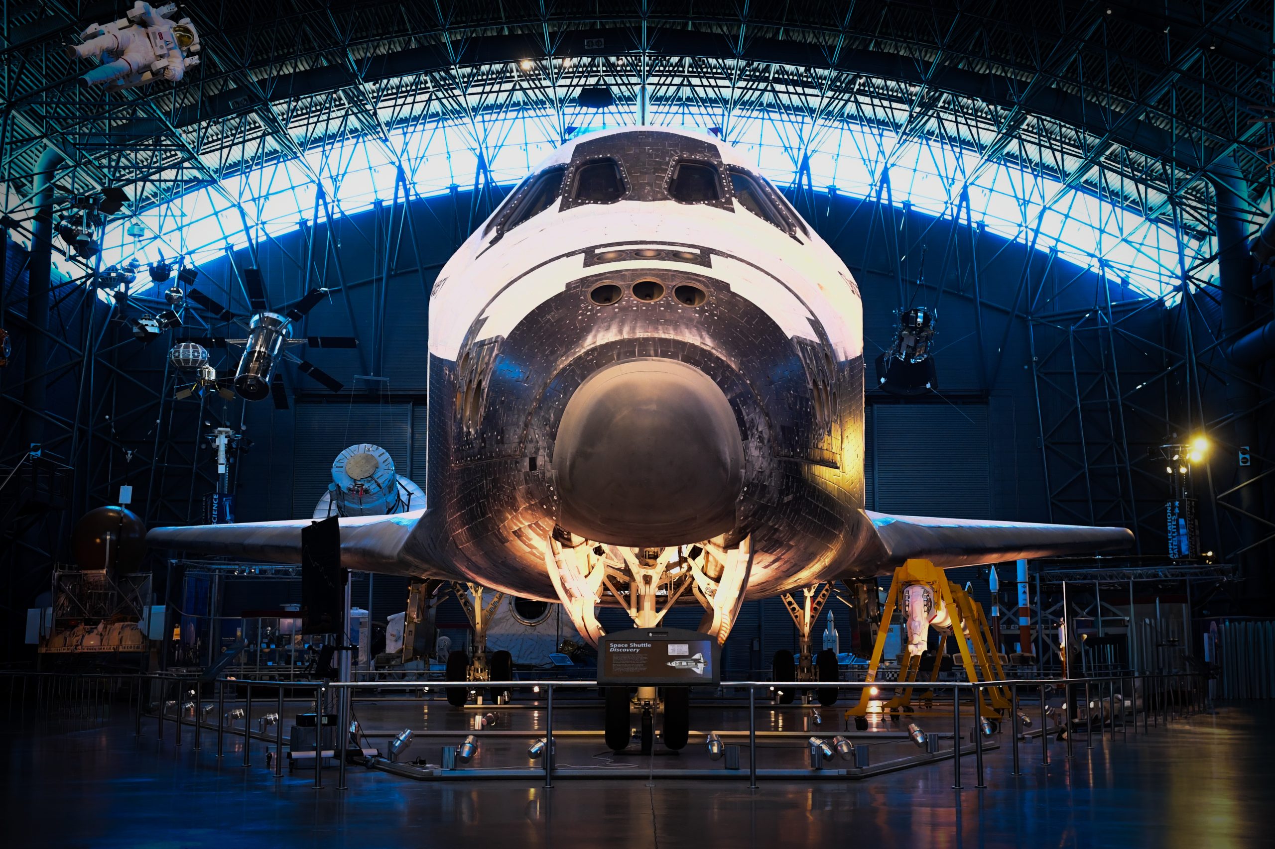 Space Shuttle Discovery on display in a large museum hangar with blue lighting and scaffolding overhead
