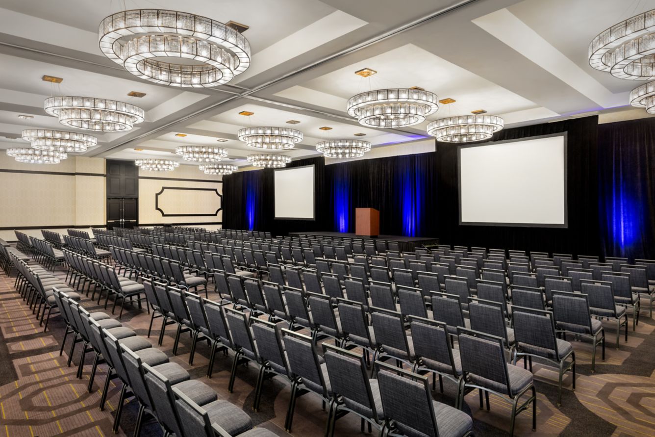 si-laxsi-ballroom-29527_Classic-Hor | ISDC Empty conference hall set up for a presentation with rows of gray chairs facing a stage, two projection screens, and a wooden podium beneath black curtains.